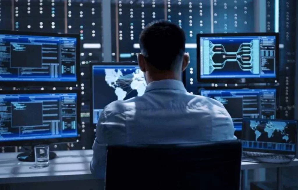 A man in front of a table full of screens, in a room full of glowing servers